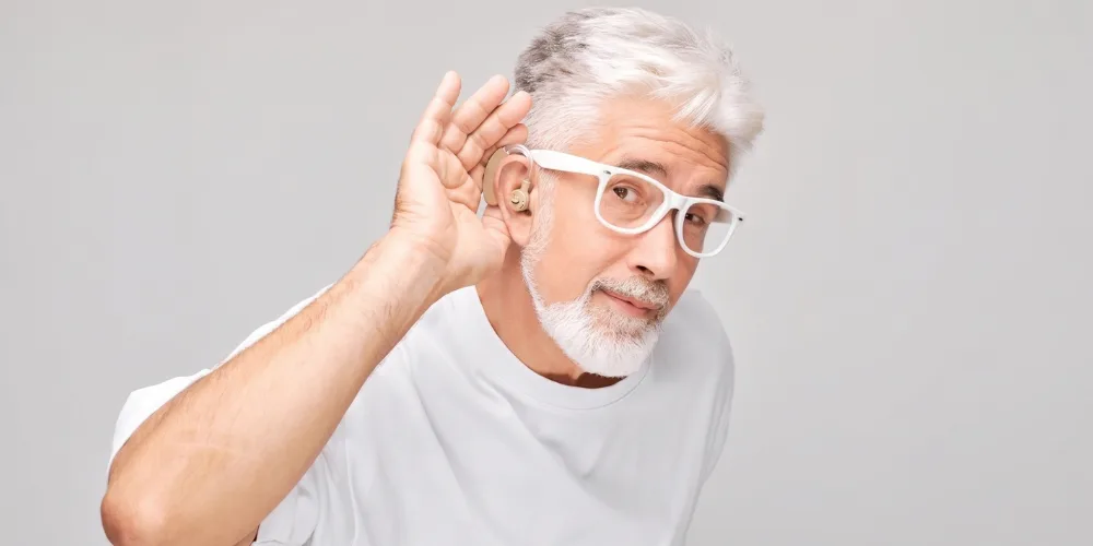 Hombre mayor con lentes blancos y barba usando un audífono retroauricular mientras se lleva la mano a la oreja en la calle.